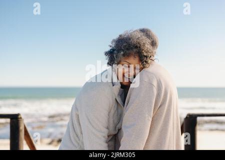 Glückliche ältere Frau, die am Meer auf die Schulter ihres Mannes gelehnt lachte. Ein romantisches, älteres Paar genießt danach einen erfrischenden Badeurlaub Stockfoto