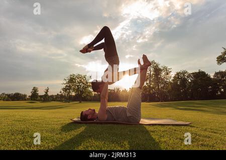 Gesunder Mann auf Gras liegend und balancierende Frau. Paar machen akrobatische Yoga-Übungen im Park Stockfoto