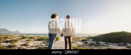 Panoramablick auf ein glückliches Seniorenpaar, das sich die Hände hält, während man am Strand eine Fußgängerbrücke hinuntergeht. Romantisches, älteres Paar, das eine erfrischende Saison nimmt Stockfoto