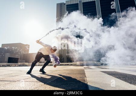 Sportlicher Mann, der Parkour und Freilauf trainiert, indem er mit einer Rauchgranate einen Flip macht. Free Runner in Aktion über städtischen Hintergrund. Stockfoto