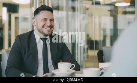 Optimistischer kaukasischer Geschäftsmann lächelt, gestikuliert und freut sich über Geschäftsgewinne, während er während des Mittagessens mit seinem Kollegen im Mod Cafe sprach. Kaffeetassen stehen auf dem Tisch Stockfoto