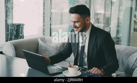 Fröhlicher bärtiger kaukasischer Geschäftsmann in formeller Kleidung, der mit seiner Familie über einen Laptop plaudert und in einem stilvollen Café fröhlich mit der Hand winkt. Er gestikuliert optimistisch und fröhlich. Stockfoto