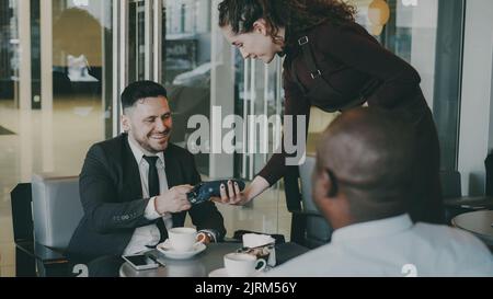 Erfolgreicher kaukasischer Geschäftsmann lächelt, sitzt mit afroamerikanischem Investor und zahlt Online-Rechnung mit seinem Smartphone in einem modernen Café. Glückliche junge Kellnerin mit ihrem tragbaren Geldautomaten. Stockfoto
