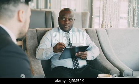 afroamerikanischer HR-Manager, der ein Vorstellungsgespräch mit einem jungen Mann im Anzug führt und sich seine Bewerbung in einem modernen Café im Haus anschaut Stockfoto