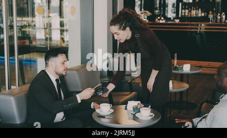 Glücklicher kaukasischer Geschäftsmann lächelt, sitzt mit seinem afroamerikanischen Partner und zahlt online Rechnung mit seinem Smartphone in einem modernen Café. Optimistische junge Kellnerin benutzt ihren tragbaren Geldautomaten. Stockfoto