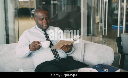 Erfolgreicher afroamerikanischer Geschäftsmann in formeller Kleidung, der Buch liest, während er in der Mittagspause im glasigen Café Kaffee trinkt Stockfoto