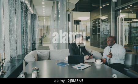 Zwei optimistische männliche Unternehmer in formeller Kleidung diskutieren im Café über ihr Startup-Projekt. Afroamerikanische und kaukasische Geschäftsleute sprechen und sitzen am Tisch mit Laptop und Charts Stockfoto