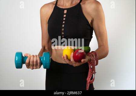 Eine Frau in Sportbekleidung hält Gemüse und Obst, Kurzhanteln, Maßband - Diät, Trainer Ernährungsberater Stockfoto