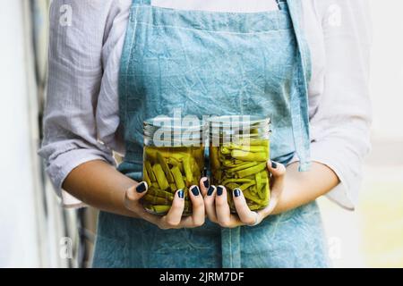 Junge Frau in einer Leinenschürze mit zwei Einmachgläsern, die mit frischen, selbst angebauten grünen Bohnen gefüllt sind. Selektiver Fokus unscharfer Hintergrund. Stockfoto