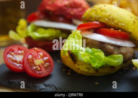 Nahaufnahme von Hamburgern mit Tomaten, Zwiebelringen und Salat und Brote in roten und gelben Farben, alles auf einem schwarzen Schiefertisch Stockfoto