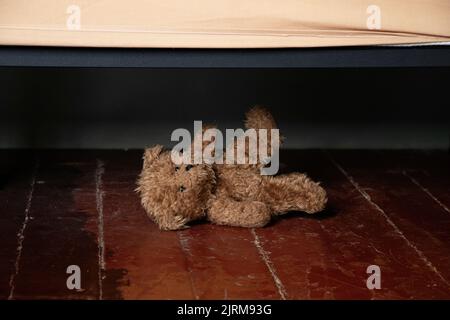 Neben dem Bett auf dem Boden im Schlafzimmer des Hauses liegt ein Teddybär, ein Teddybär unter dem Bett auf dem Boden im Dunkeln Stockfoto