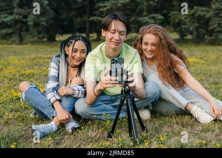 Gruppe junger Influencer Teenager sitzen auf dem Rasen im Park und machen ein Live-Video im Internet. Freundschaft und neue Technologiekonzepte in einem Stockfoto