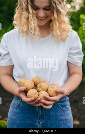 Glückliche Bäuerin mit lockigen Haaren lächelt fröhlich, nachdem sie frische Kartoffeln aus ihrem Bio-Garten geerntet hat. Nahaufnahme Porträt. Gesunder Lebensstil. Stockfoto