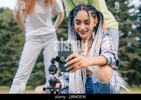 Frau nutzt die Video-Sharing-App auf dem Mobiltelefon, während sie mit ihren Freunden im Park steht. Moderne Kommunikation, Social Media und Gadgets. Stockfoto