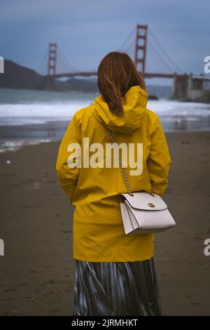 Eine Frau in einer gelben Regenjacke, die an einem Strand steht und auf die Golden Gate Bridge in San Francisco blickt Stockfoto