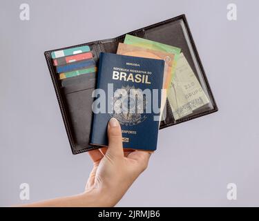 Brasilianischer Reisepass in einer Hand mit Geldbörse, Kreditkarten und Euro-Banknoten auf weißem Hintergrund. Stockfoto