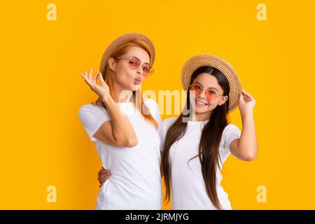 Mutter und Tochter im Teenageralter umarmen und kuscheln sich lieblich, tragen T-Shirt Strohhut und Sonnenbrille, isoliert auf hellgelbem Hintergrund. Sommer Stockfoto