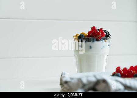 Joghurt, Haferflocken-Frühstücksflocken und frische Sommerbeeren im Glas auf dem weißen Tisch. Rote Johannisbeeren, Blaubeeren, Brombeeren auf weißem Teller, Stockfoto
