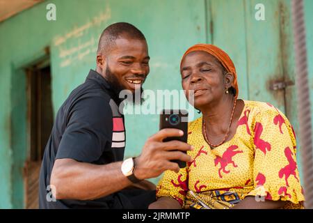 Lächelnder afrikanischer Mann zeigt einer alten afrikanischen Frau Inhalte auf seinem Telefon Stockfoto