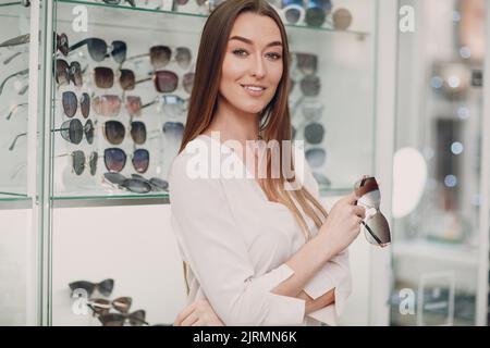 Nahaufnahme einer wunderschönen jungen Frau, die lächelt und an der Optikerecke im Einkaufszentrum Gläser pflückt und auswählt. Glückliche, schöne Frau, die beim Optiker Brillen kauft. Stockfoto