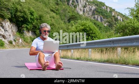 Grauhaariger älterer Mann, Freiberufler mit Bart in Sonnenbrille, der an einem Laptop auf Natur, Straße und Bergen arbeitet. Ein verrückter und außergewöhnlicher alter Mann sitzt im Sommer auf einer Yogamatte Stockfoto