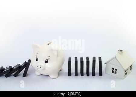 Domino-Fliesen, die von einem weißen Sparschwein unterstützt werden und ein weißes Modellhaus schützen. Isurance kreatives Konzept, Nahaufnahme. Stockfoto