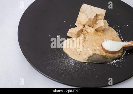 Auflösen von frischer Hefe mit Zucker auf einem schwarzen Teller. Nahaufnahme der Seitenansicht Stockfoto