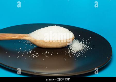 Holzlöffel voller Kristallzucker auf einer schwarzen Keramikplatte, Nahaufnahme von der Seite Stockfoto