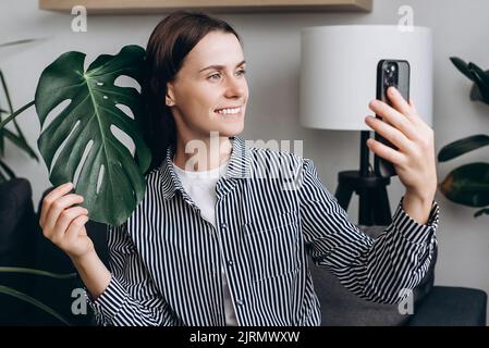 Lächelnde Gärtnerin, die nach Transplantationen von Zimmerpflanzen angenehme Gespräche mit einer mobilen Webcam-Videoanruf-Anwendung genießt. Konzept Werk ca. Stockfoto