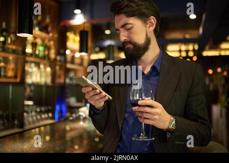 Unglücklicher Mann mit einem Glas Rotwein, der Nachrichten in seinem Telefon las Stockfoto