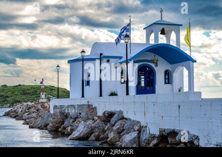 Kleine Kapelle mit weißen Dächern auf Klippe über Meer und Kleine Bucht unter einem dramatischen Himmel auf einer griechischen Insel Stockfoto