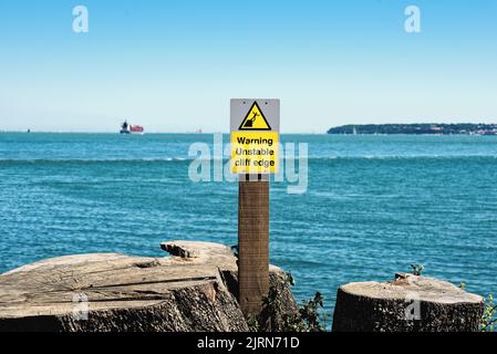 Ein gelbes Warnschild warnt die Öffentlichkeit vor den Gefahren einer instabilen Klippenkante im Lepe Country Park mit dem Solent im Hintergrund von Hampshire UK Stockfoto