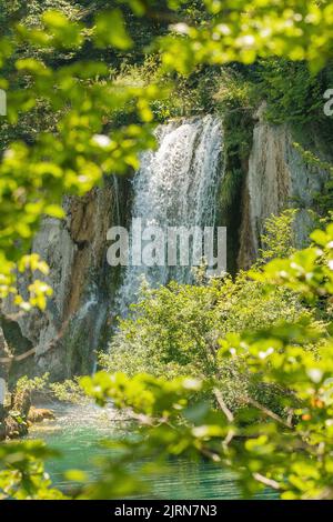 Der Wasserfall fließt bei Sonnenlicht von einer felsigen Klippe in klares türkisfarbenes Wasser. Atemberaubende Aussicht auf die Aquamarin-Kaskade im Nationalpark auf den Plitvicer Seen Stockfoto