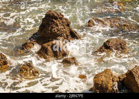 Kleine Meereswellen, die bei Flut bei hellem Sonnenlicht auf der flachen Küste von Felsen abstürzen. Transparente Wellen erzeugen Schaumstoffe, die auf Steinen Rollen Stockfoto