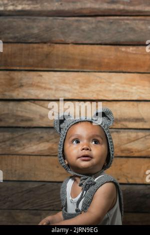 Schöne Latina mit brauner Haut, mit Overalls und einer Mütze mit Mausohren, auf einem hölzernen Hintergrund. Neugierig nach oben schauend, auf der Suche nach ihr Stockfoto