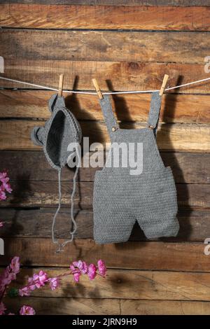 Babykleidung, die an einem Wäscheleine-Seil hängt, das mit Holzhaken, Overalls und Hut mit in der Hand gehäkelten Technik gestrickten Ohren auf einem hölzernen Bac gebunden ist Stockfoto