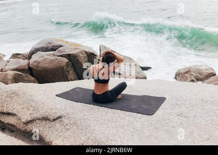 Rückansicht einer jungen Frau, die auf einer Matte sitzt und ihren Hals am Meer aufwärmt. Frau, die Yoga macht und auf Wellen schaut. Stockfoto