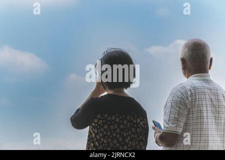Rückansicht des Glücks Asiatisches älteres Paar mit Mobiltelefon mit Wolke und Himmel im Hintergrund. Das ältere Paar ruht die Augen, während es auf die Schönheit schaut Stockfoto