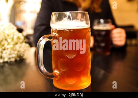 Defokussieren Sie den Becher mit leichtem Bier. Nahaufnahme von zwei Händen klirrenden Bier stein Gläser. Zwei Freunde trinken Bier im Pub. Dating. Nicht fokussiert. Stockfoto