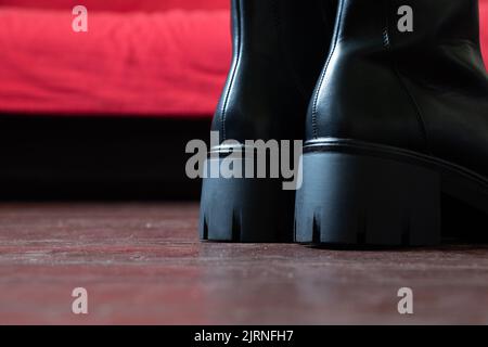 Mädchen in hohen schwarzen Stiefeln auf dem alten schmutzigen Boden zu Hause, Damenschuhe, Füße in Schuhen auf dem Boden, Schuhe auf dem Holzboden Stockfoto