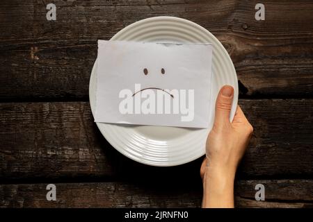 Auf einer Karte, die in den Händen eines Mädchens auf dem Tisch auf einem weißen Teller liegt, wird ein trauriges Emoticon gezeichnet, eine schlechte Laune und eine Krise, Armut und Traurigkeit Stockfoto