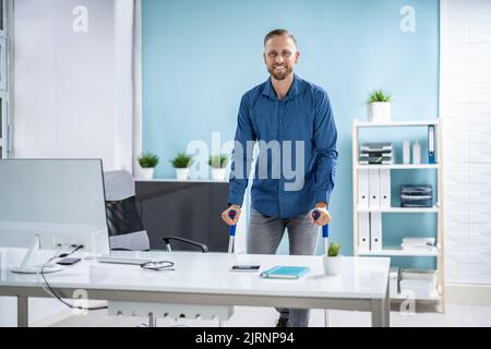 Arbeiter Mit Krücken Am Arbeitsplatz Oder Im Büro. Rehabilitationsleistungen Stockfoto