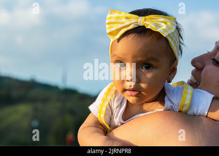 Schönes braunhäutig geblieenes Baby auf der Schulter ihrer Mutter, das sehr neugierig nach hinten schaut. Junge Latina-Mutter lächelt fröhlich, während sie stolz anschaut Stockfoto