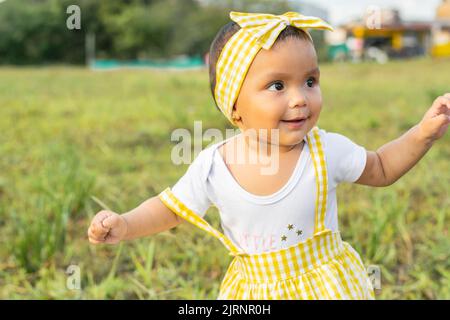 Kleines lateinisches Mädchen, das auf einem grasbewachsenen Feld steht, lächelt und sieht sehr glücklich aus. Schöner Sommertag in der kolumbianischen Kaffeeregion. Baby mit brauner Haut Stockfoto