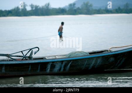 Thailändisches kleines Fischerboot, das am Strand festgemacht ist und die Fischer sich morgens im Hintergrund darauf vorbereiten, angeln zu gehen. Das Leben im traditionellen Thai Coun Stockfoto