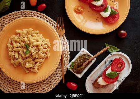 Italienische Pasta-Fusilli, Caprese-Salat - Tomaten, Basilikum, Mozzarella und Pesto in niedriger Tonart. Brotvorspeise mit Caprese-Salat. Blick von oben. Italienisches Abendessen Stockfoto