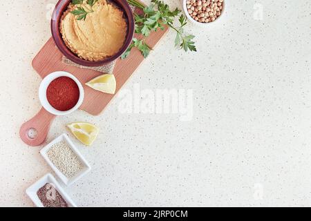 Cremiger Hummus auf dem Schneidebrett mit Petersilie, Zitrone, Paprika, römischen Samen, Kichererbsen, Sesamzutaten. Gesunde vegetarische natürliche rustikale Nahrung. Lebensmittel von oben, Kopierbereich. Stockfoto