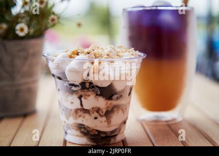 Leichte natürliche Portion Nüsse Dessert in einer Einweg-Tasse. Lila und gelber Cocktail mit Strohschilf. Anchan-Blautee mit Antioxidantien und Nootrepien auf der Terrasse in der Nähe von Kamillenblüten. Stockfoto