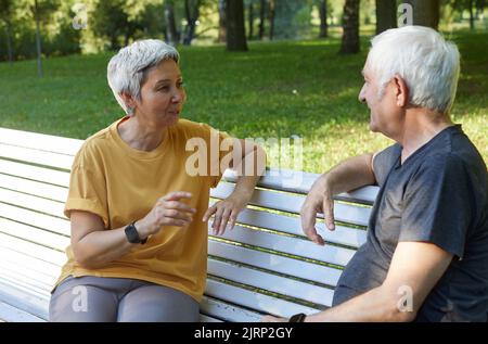Attraktives älteres Paar, das auf der Bank im Sommerpark sitzt, sportliche Kleidung trägt, sich morgens im Freien unterhalten kann. Kommunikation, Schaden Stockfoto