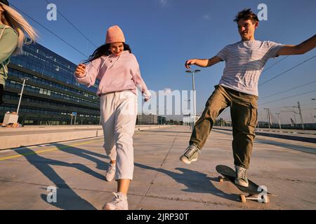 Drei aufgeregte Teenager verbringen Zeit im städtischen Außenbereich. Sie laufen und Skateboarden. Stockfoto